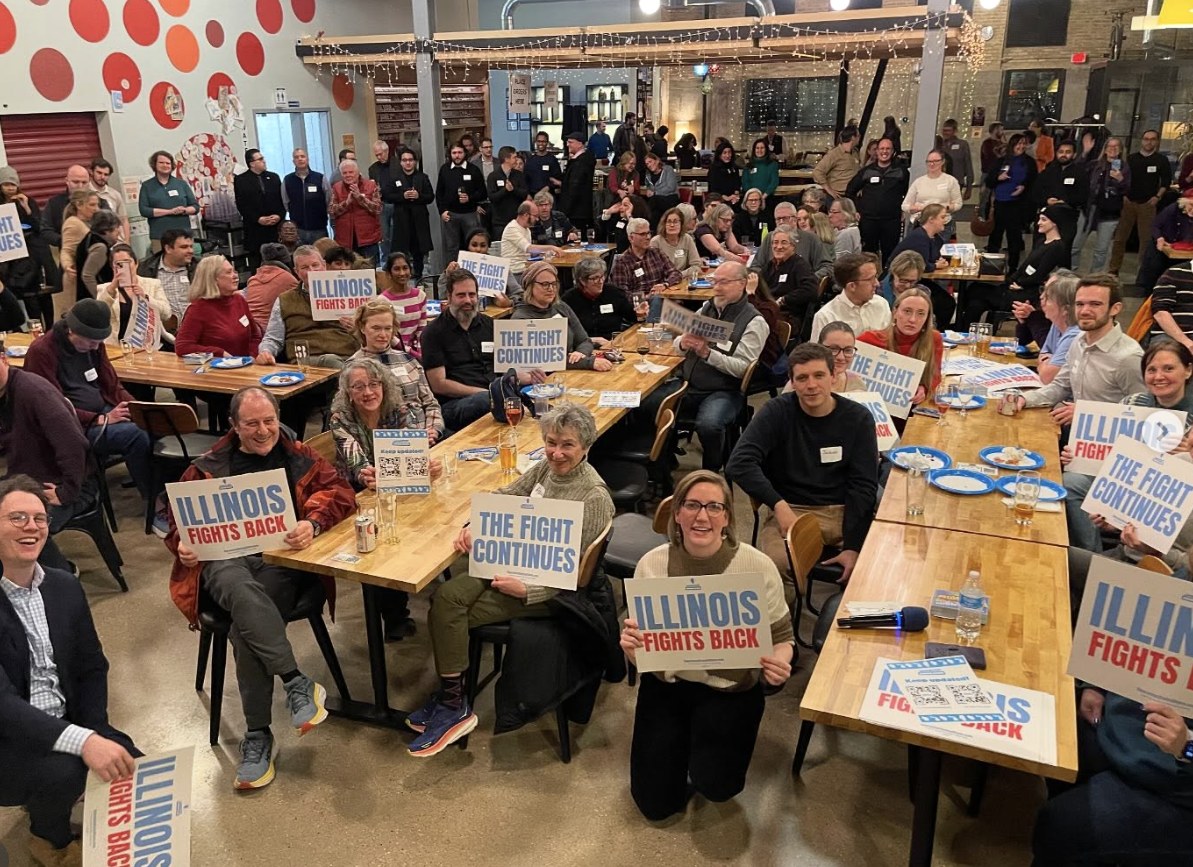 A packed room of volunteers holding Illinois Fights Back signs at the 2026 midterm kickoff